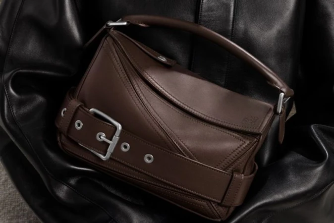 Studio shot of a brown leather LOEWE handbag with a silver buckle and strap, draped over a black leather jacket.