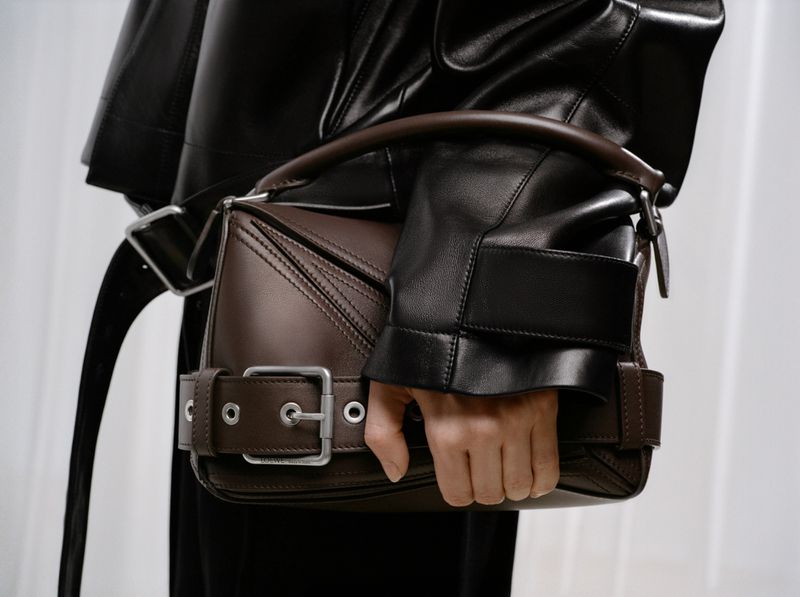 Close-up of a brown leather handbag with a silver buckle, held by an adult female wearing a dark leather jacket against a white background.