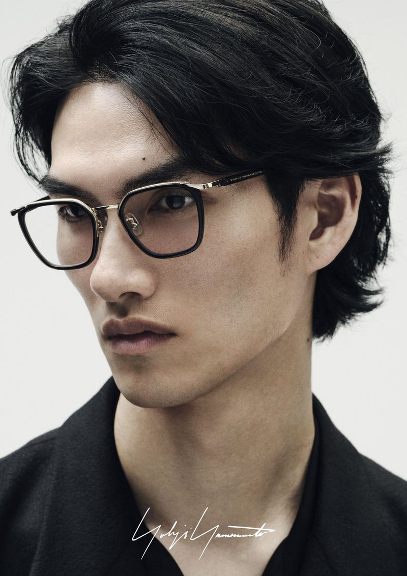 Close-up of a young Asian man wearing black-and-gold eyeglasses and a black jacket against a neutral background.