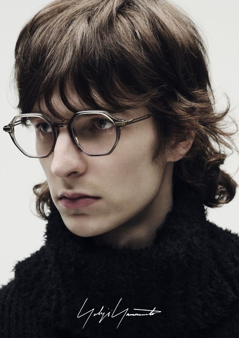 Portrait of a young adult with long brown hair and octagonal glasses wearing a dark sweater against a neutral background.