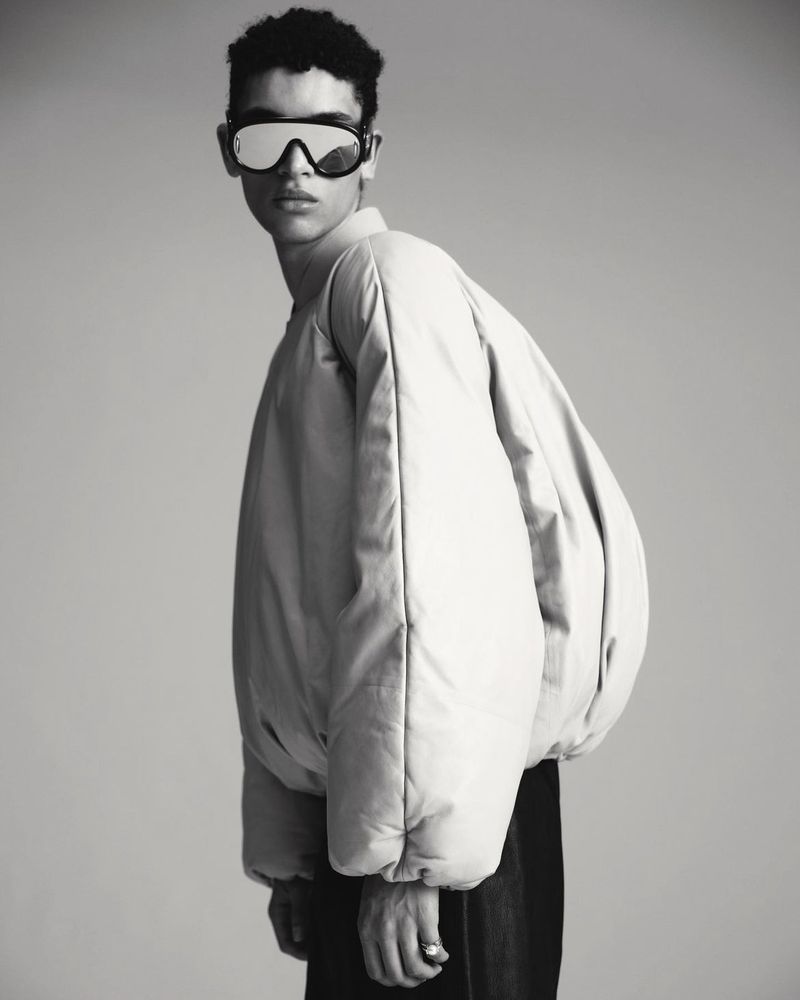 Monochrome portrait of a person with dark, curly hair wearing a puffy jacket and reflective goggles against a grey background.