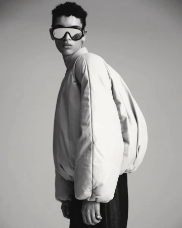 Monochrome portrait of a person with dark, curly hair wearing a puffy jacket and reflective goggles against a grey background.