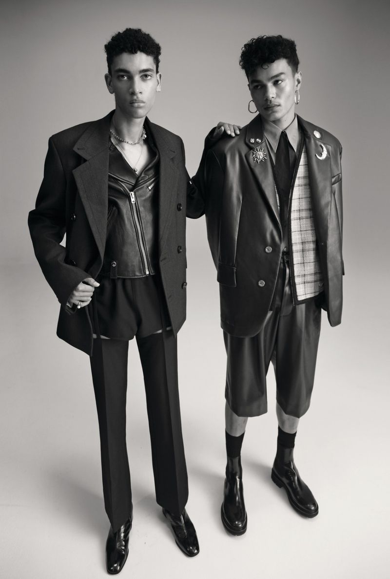 Two male models pose in a photo studio wearing black outfits and shiny black dress shoes, conveying a high-fashion editorial vibe.