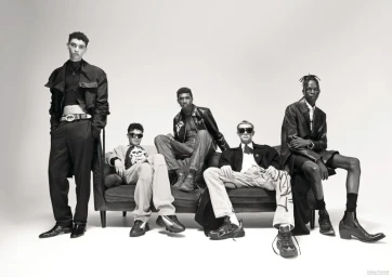 Five male models posing in contemporary black streetwear on and around a dark couch against a plain white studio backdrop.