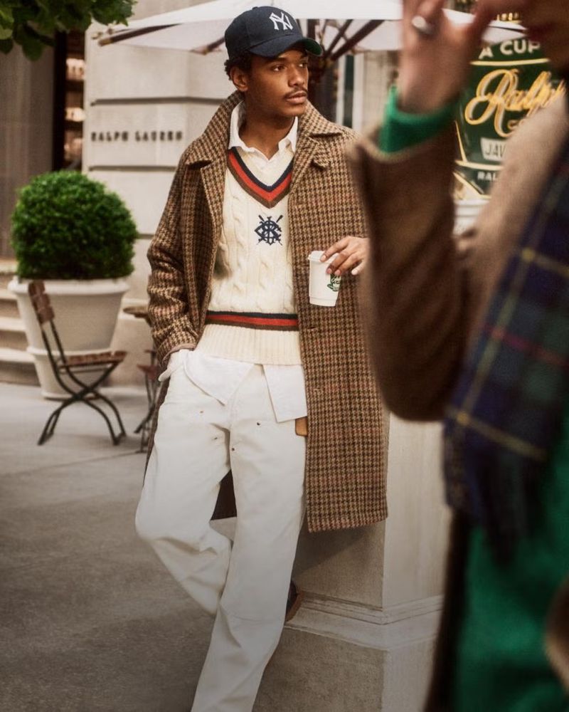 A stylish adult male with a baseball cap and coat stands on a sidewalk with a coffee cup in hand in an outdoor urban environment.