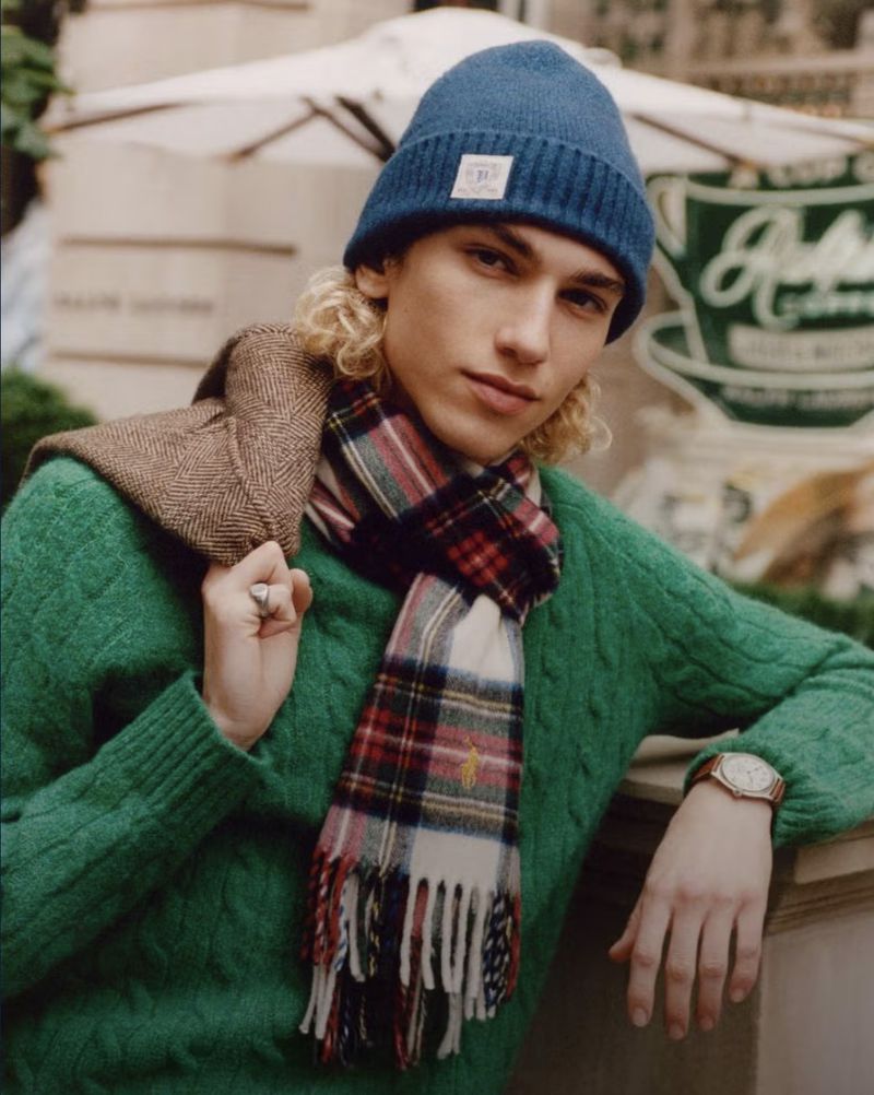 A young adult male with blond curly hair models a blue beanie, plaid scarf, and green sweater, posing in front of a cafe sign.