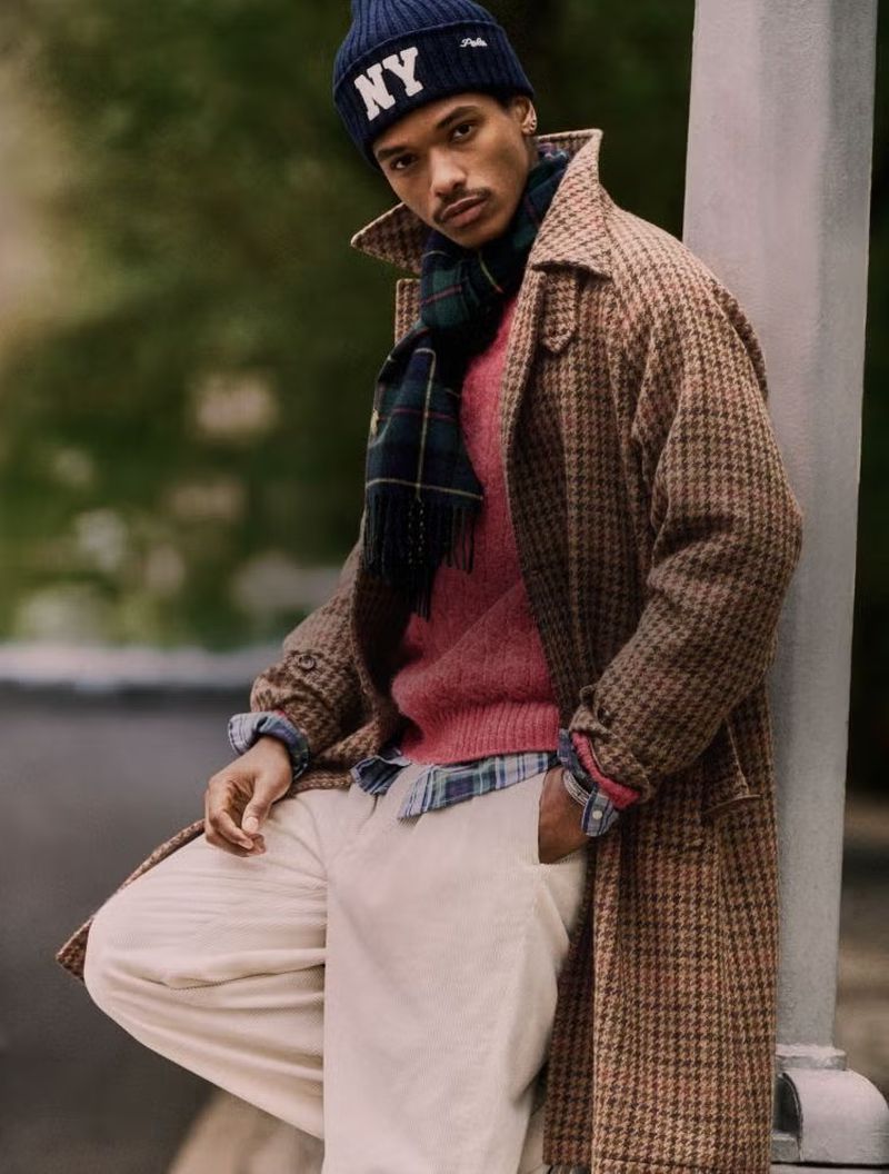 Man leans against post wearing beanie, scarf, overcoat, pink sweater and pants in an outdoor setting. Fall or winter fashion.