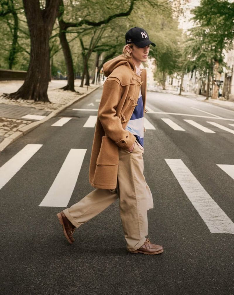 An adult male is walking across a crosswalk wearing a tan duffle coat, beige pants, a striped shirt, brown shoes, and a baseball cap.