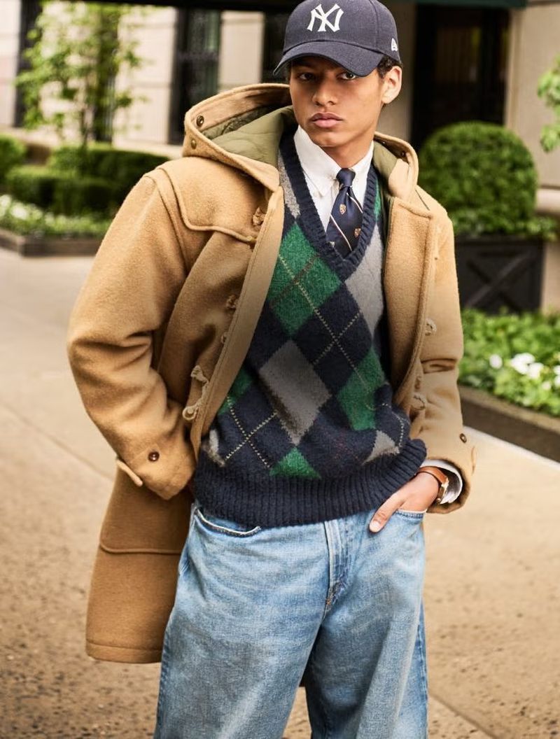 Man in coat, argyle sweater, jeans, and baseball cap in an outdoor setting, showcasing fashion and style in natural lighting.