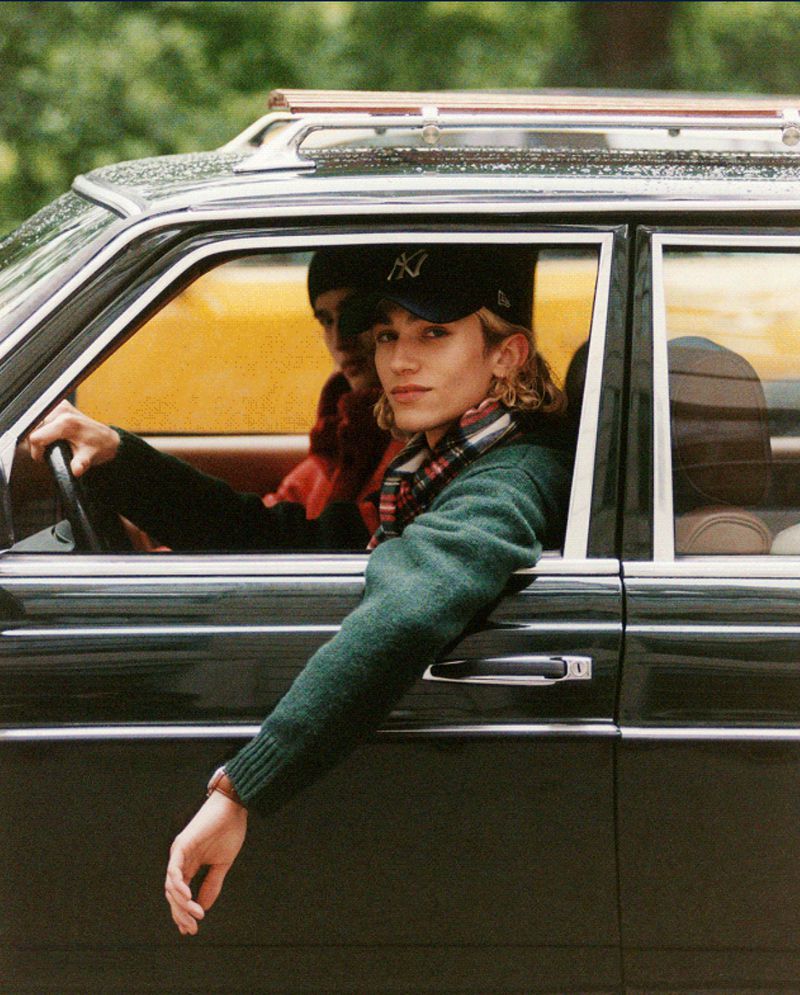 Young man in a black car wearing a baseball cap and a green sweater looking towards the camera, with another passenger in the back seat.