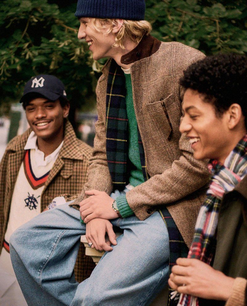 Three smiling young men wearing autumn fashion including coats, sweaters, jeans, and caps are posing in an outdoor setting.