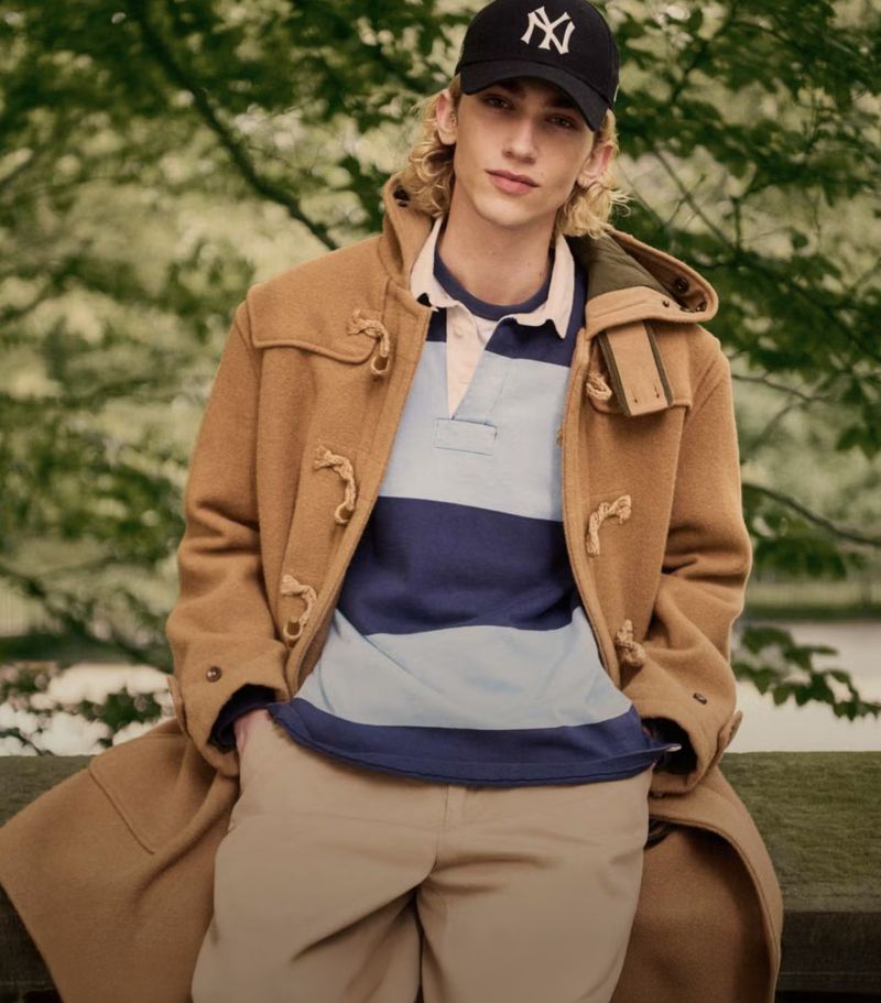 Man in a tan duffel coat and baseball cap poses outdoors. He is wearing a blue and white striped rugby shirt and khaki pants.