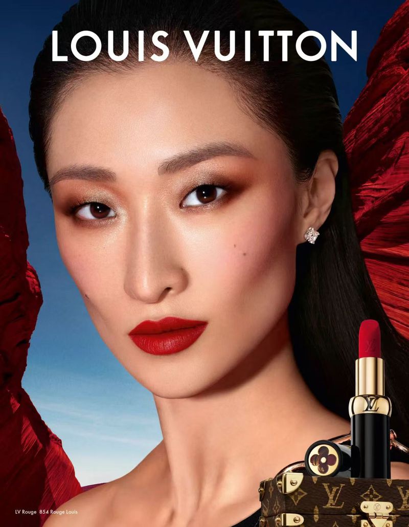 Portrait of an Asian woman with red lipstick and eye shadow, wearing earrings and posing with Louis Vuitton lipstick and case.