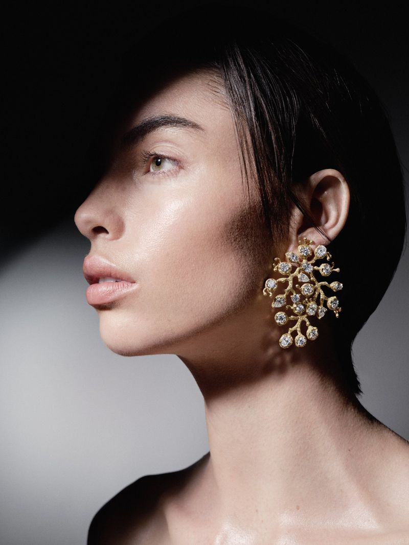 Profile shot of a woman with short hair wearing a large gold and diamond statement earring in a studio setting with dark lighting.