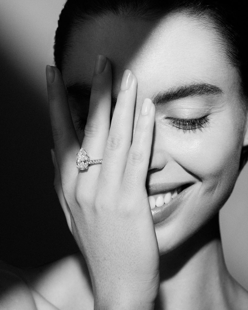 Close-up portrait of a smiling adult woman's face with hand near her face, wearing a diamond engagement ring, in black and white.