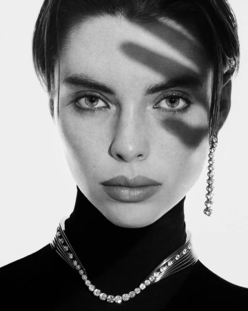 Close-up black and white portrait of a woman wearing a black turtleneck, diamond necklace, and earring, with a shadow on her face.