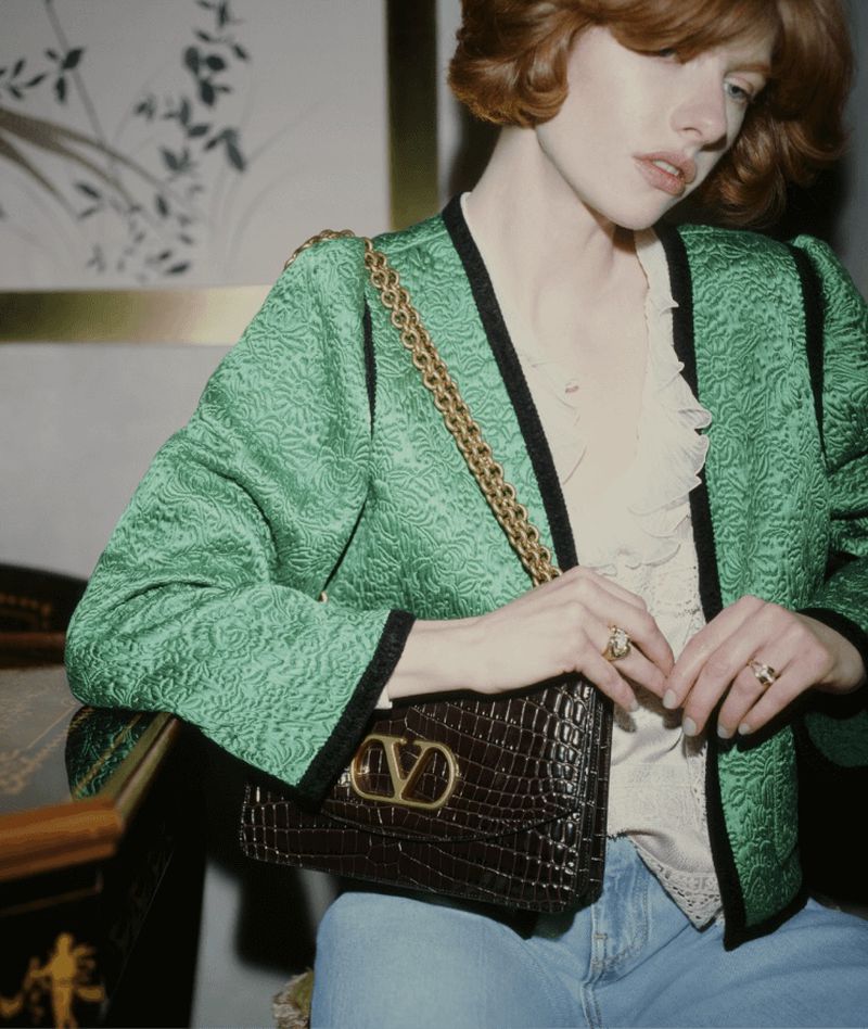 Portrait of a redhead woman wearing a green patterned jacket, light blue jeans, and holding a brown crocodile leather handbag with a gold chain.