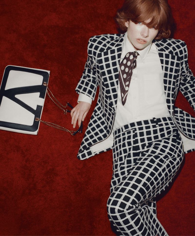 A woman in a checkered suit lies on a red surface next to a white bag with a black 'V' on it, showcasing fashion apparel.