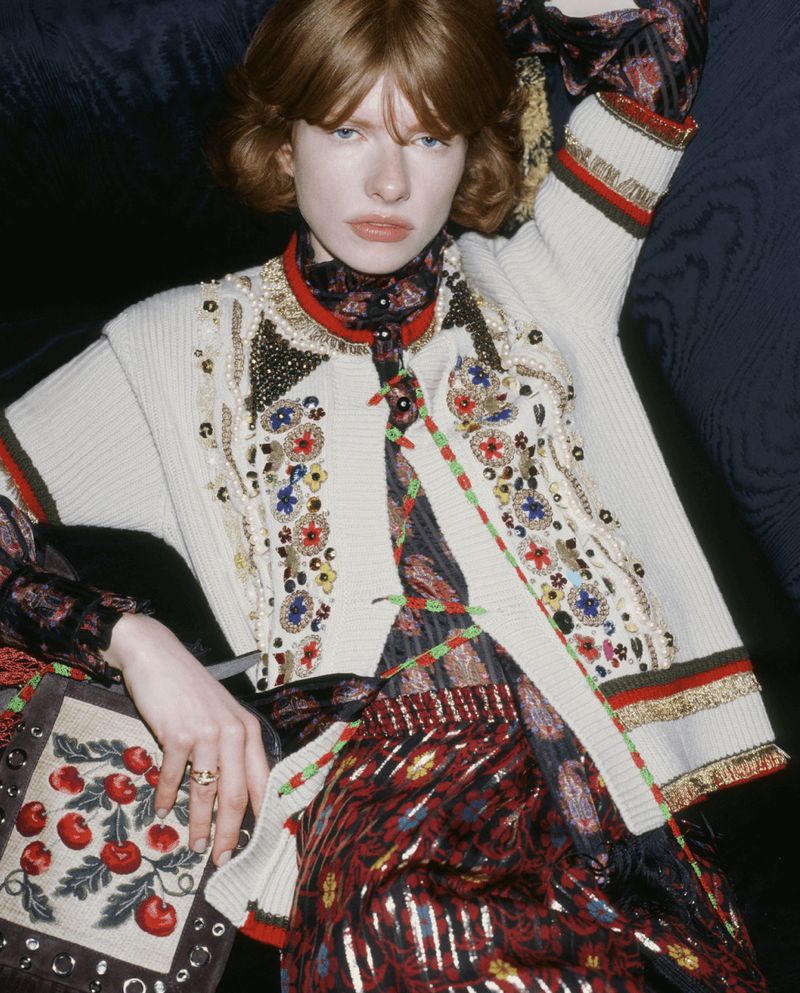 A woman with fair skin and reddish-brown curly hair poses indoors, wearing a cream embroidered cardigan over a patterned dress. 