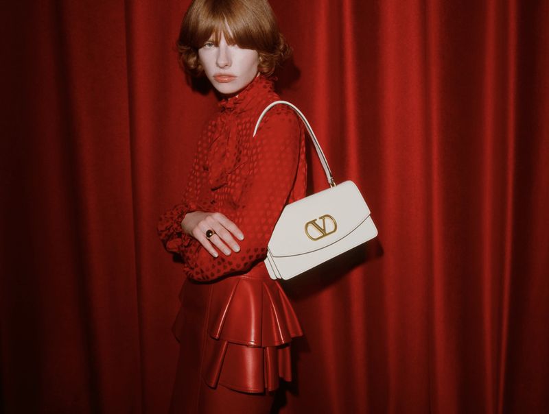 Fashion model posing with a Valentino purse, wearing a red tiered leather skirt and a patterned red top against a red backdrop.
