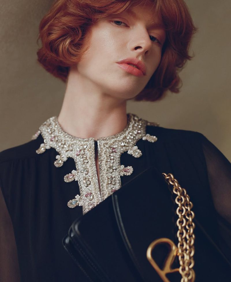 Close-up of a red-haired woman in an embellished collar dress holding a black bag with a gold chain in a muted indoor setting.