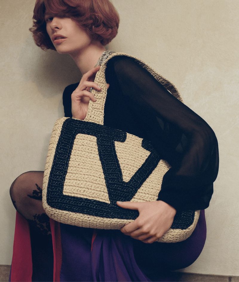 Woman poses indoors with a beige Valentino handbag featuring a large black 'V' logo, styled with a black blouse and colorful maxi skirt.