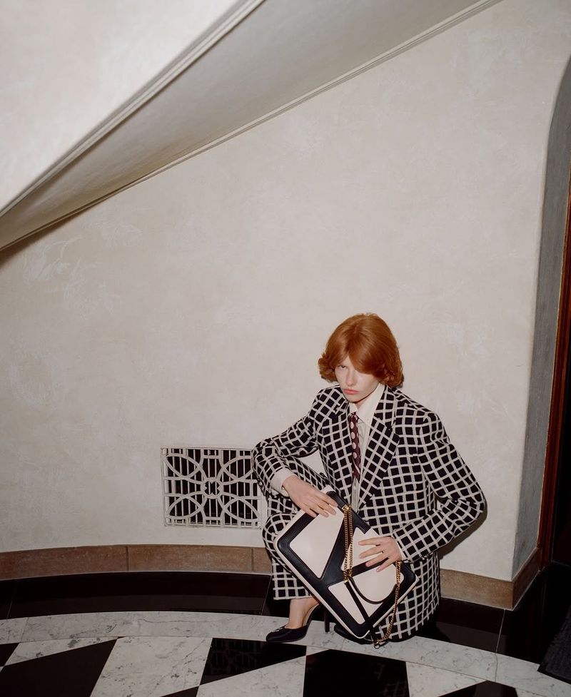 Elegant red-haired woman in checkered suit with structured handbag. Seated in interior against wall with geometric tile floor and vent.