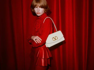 Model with red hair wearing a red blouse and skirt holding a white shoulder bag against a red curtain background.
