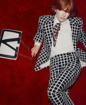 Model in a black and white grid suit and polka dot scarf lying on a plush red surface with a graphic logo handbag beside her.