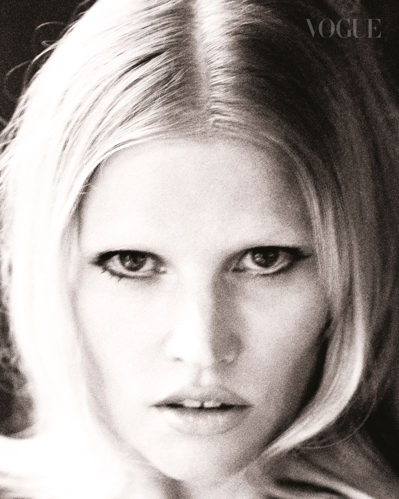Monochromatic close-up portrait of a woman with blonde hair and makeup, featuring the Vogue logo in the upper right corner.
