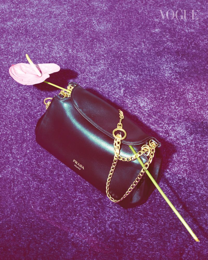 Black Prada handbag with gold chain and a pink anthurium flower against a purple textured backdrop in a studio setting for Vogue.