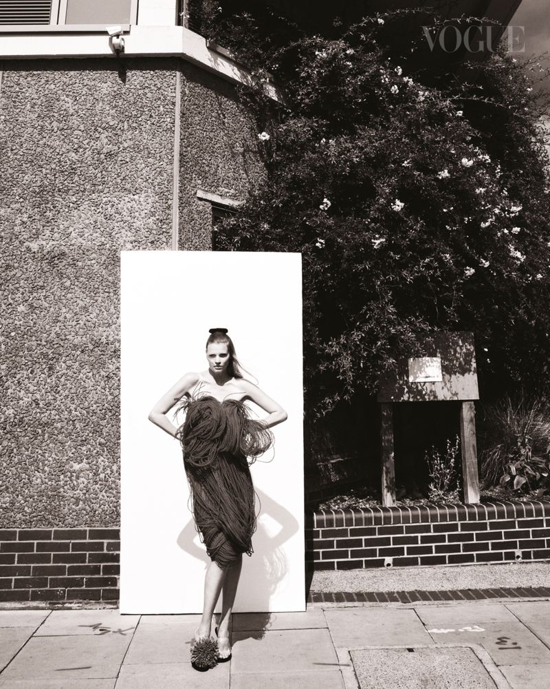Monochrome image of a woman in a fringed dress posing in front of a white board, with a building and trees in the background.