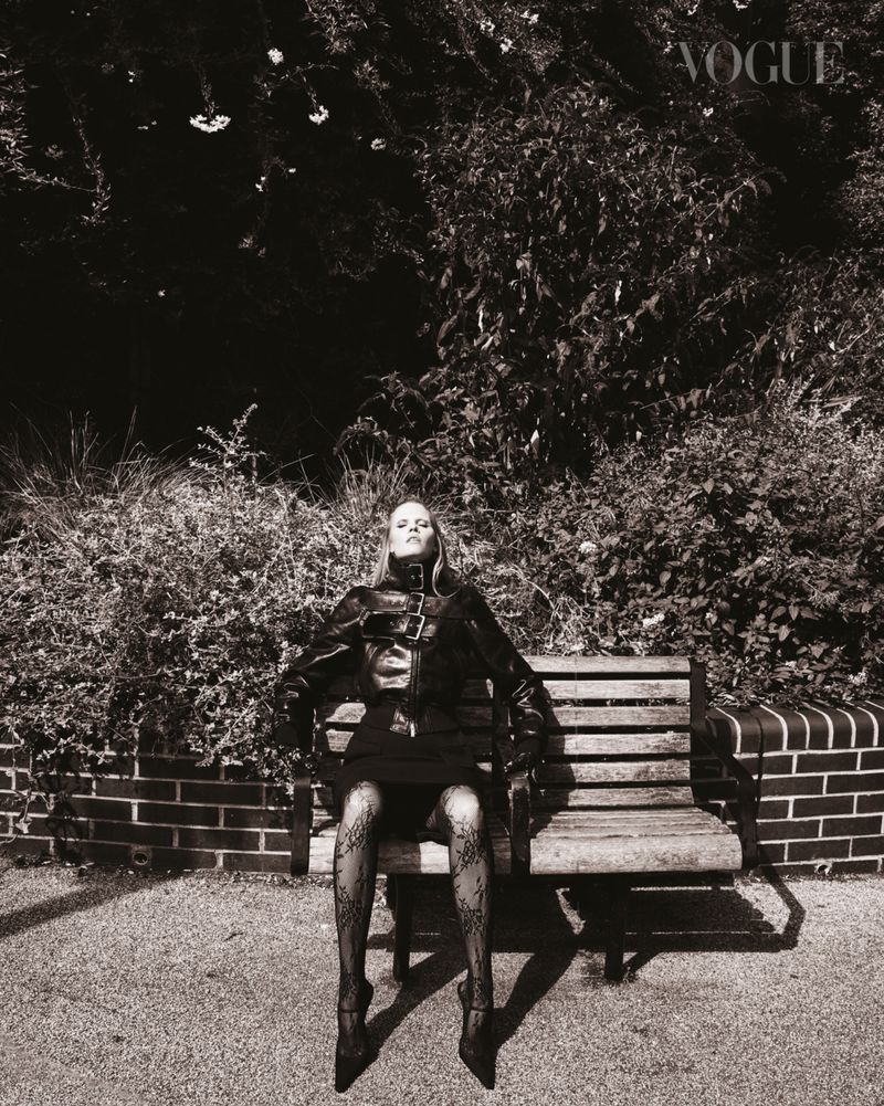Monochrome portrait of an adult woman in a jacket and short skirt, posing on a bench in front of trees and a brick wall. Vogue logo visible.
