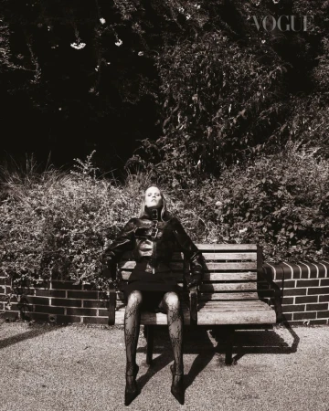 Monochrome portrait of an adult woman in a jacket and short skirt, posing on a bench in front of trees and a brick wall. Vogue logo visible.