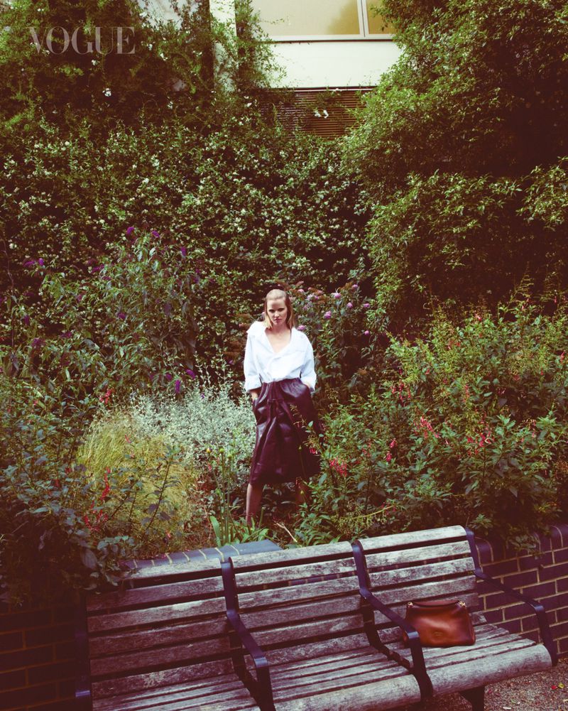 A woman stands in a lush garden setting, wearing a white button-down shirt and a dark skirt, with a wooden bench and purse in the foreground.