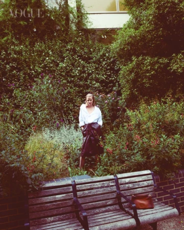 A woman stands in a lush garden setting, wearing a white button-down shirt and a dark skirt, with a wooden bench and purse in the foreground.