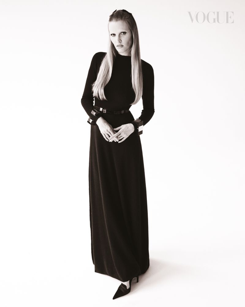 Full-length shot of a blond woman wearing a long black dress and accessories against a bright white backdrop. Fashion editorial.