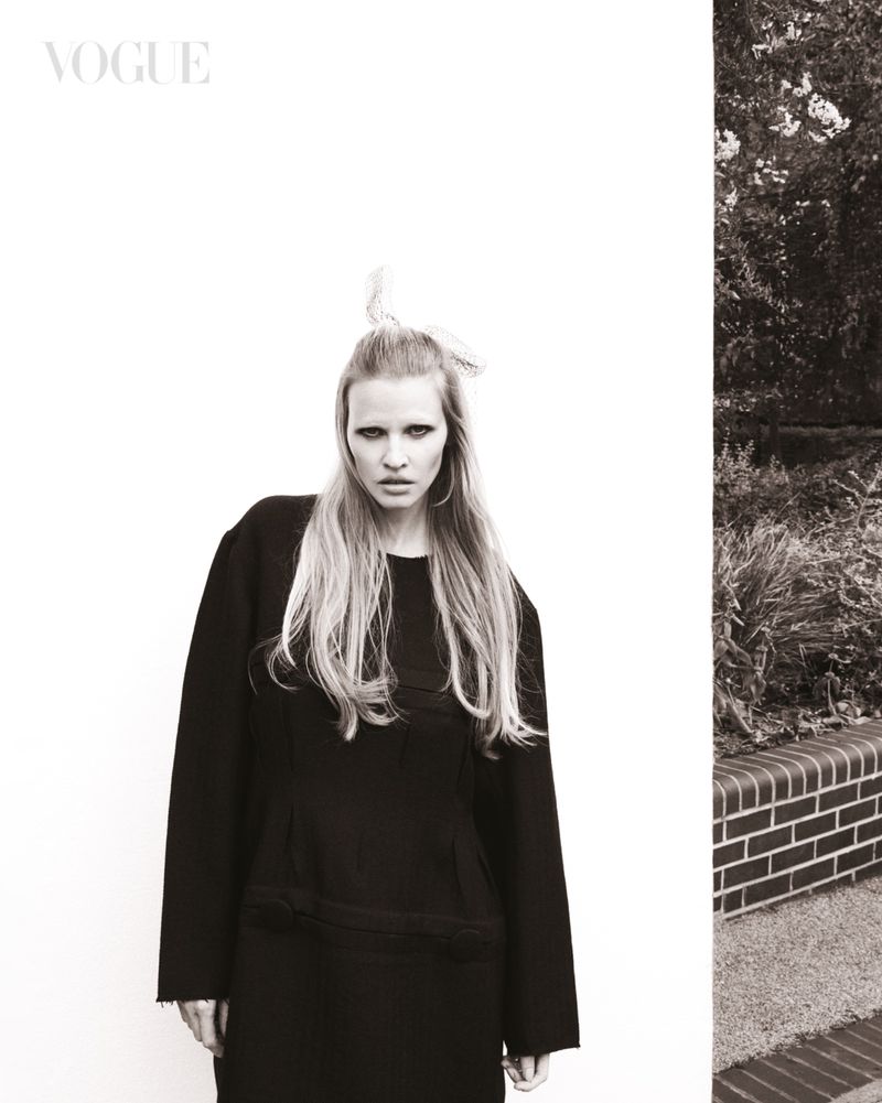 A black-and-white fashion shot featuring a slender adult female model in a dark, oversized dress, set against a white wall and garden background.