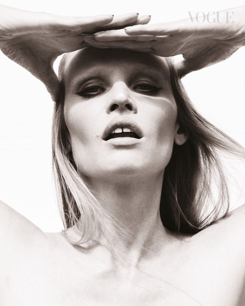 Monochrome portrait of a woman with makeup, fair skin, light hair and hands above her head in front of a bright background.
