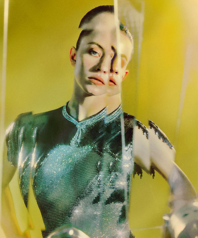 Woman posing in reflective surface with a yellow backdrop wearing a shimmering top, creating a distorted, surreal portrait.