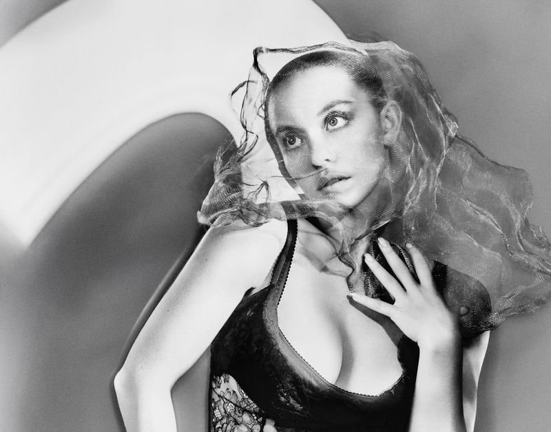 Black and white studio portrait of a young woman wearing a lace top and a net veil draped over her head and arm.