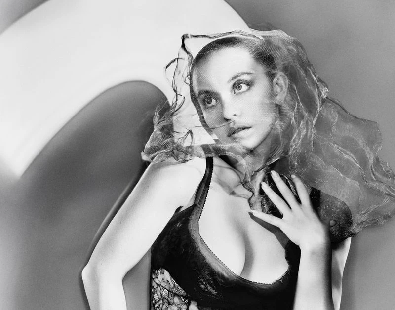 Black and white studio portrait of a young woman wearing a lace top and a net veil draped over her head and arm.