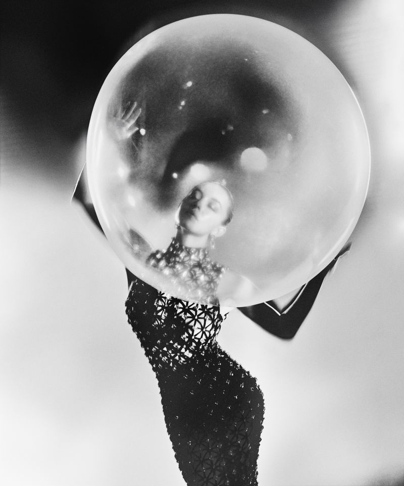 Monochrome fashion shot of a woman in a patterned gown, posing inside a large, transparent bubble against a gradient grayscale background.