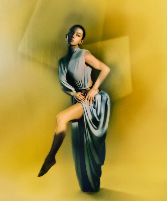 A woman in a gray dress poses with one leg raised against a yellow background with blurred square light patterns, creating an elegant portrait.