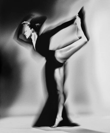 Sydney Sweeney performing a ballet dancer leg extension in a black dress in a black and white studio portrait.