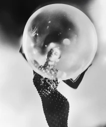 Sydney Sweeney holding a large glass sphere reflecting light while wearing a dark patterned dress in a monochrome editorial portrait.