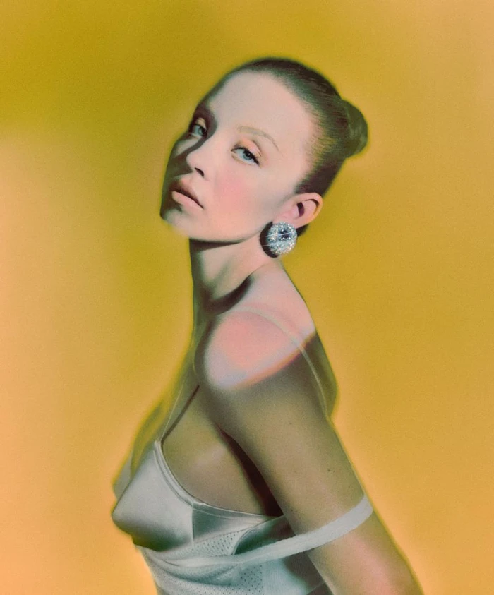 Portrait of Sydney Sweeney wearing a white top and statement earring against a warm yellow gradient background.