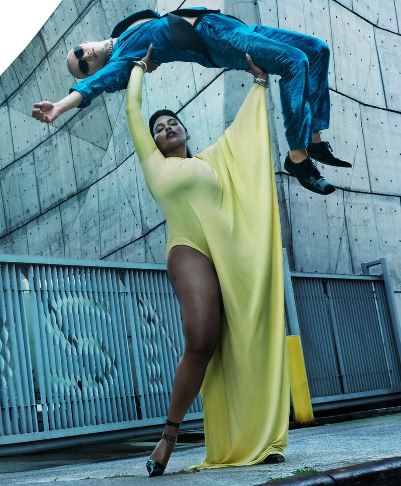 Woman in yellow leotard lifting a bald man in a blue suit against a concrete background. Fashion-forward scene with dynamic posing.