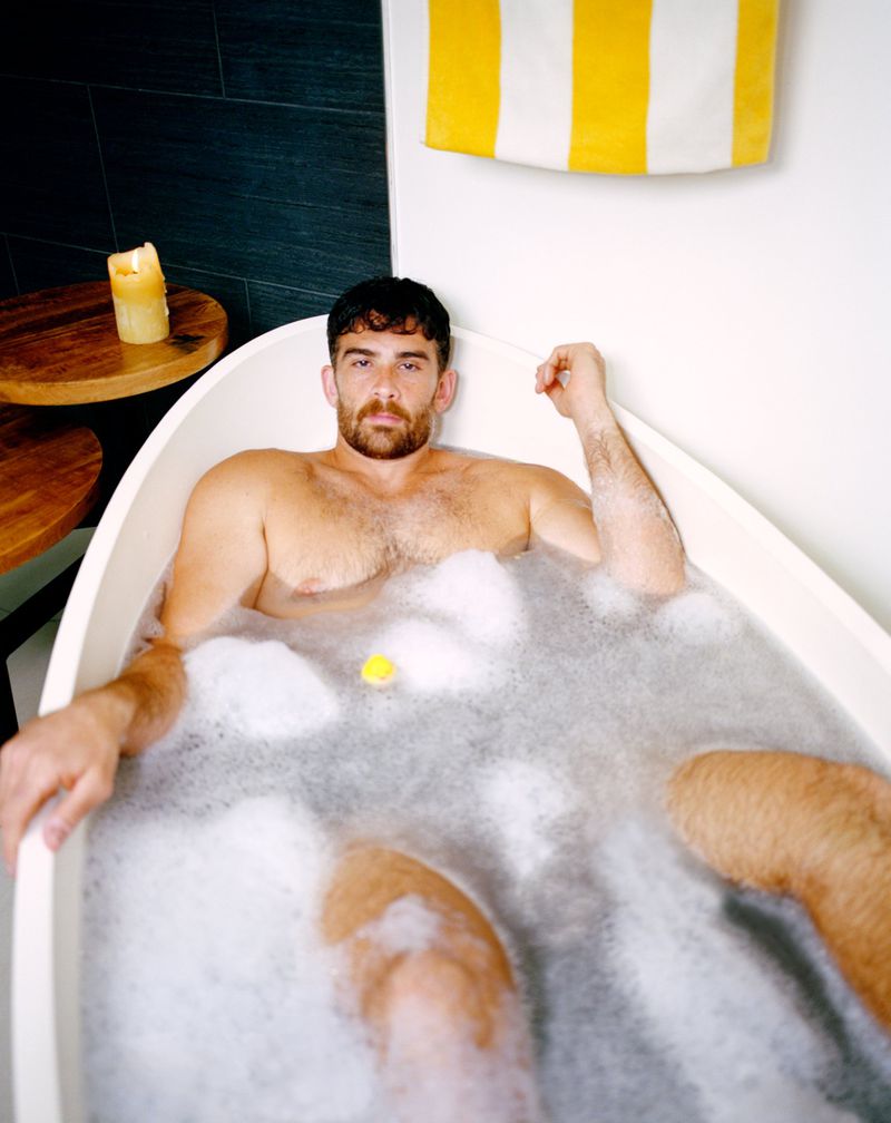 Adult male relaxing in a bubble bath with a rubber ducky and a towel. Indoor bathroom setting with white walls and tile.