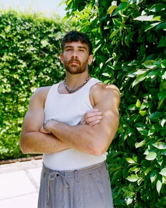 Hasan Piker wearing a white ribbed tank top and patterned trousers standing outdoors against a green leafy hedge.